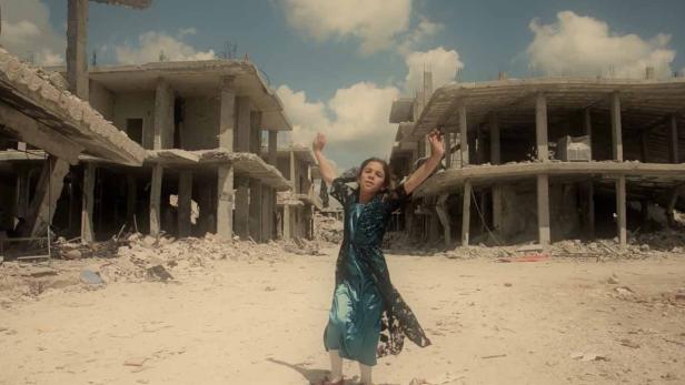 Ein junges Mädchen steht inmitten zerstörter Gebäude und hebt die Arme.