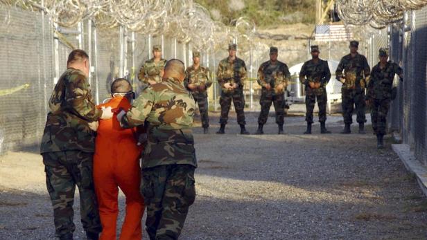 Soldaten in Tarnuniformen führen einen Gefangenen in orangefarbener Kleidung ab.