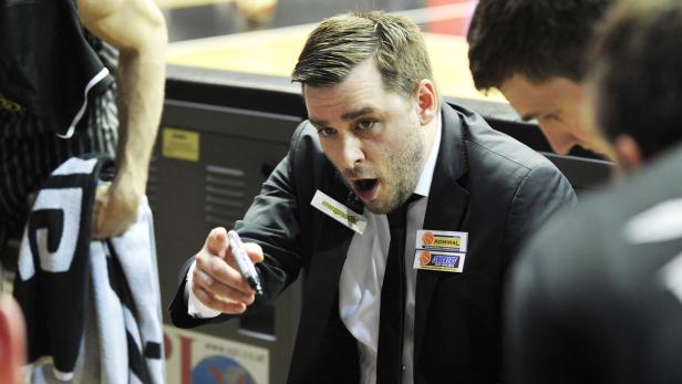 Ein Basketballtrainer in Anzug erklärt eine Spielstrategie mit einem Stift in der Hand.