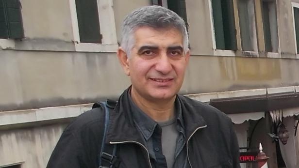 Ali Bayraktar hat ein graues Oberteil und eine schwarze Jacke an, er steht vor einer grau-braunen Hausmauer
