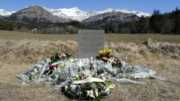 Ein Denkmal mit Blumen und Kränzen auf einer Wiese vor einer Bergkulisse.