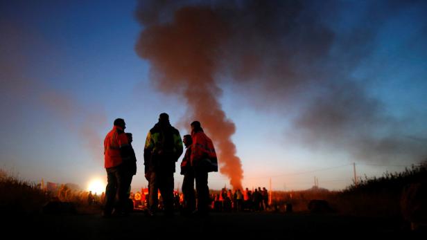 Eine Gruppe von Menschen steht vor einer Rauchsäule bei Dämmerung.