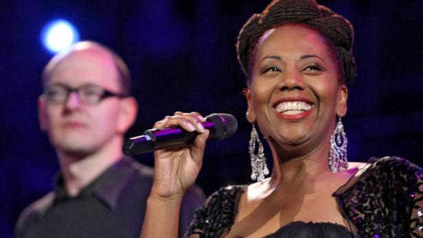 Eine lächelnde Sängerin mit Mikrofon auf der Bühne, begleitet von ihrer Band.