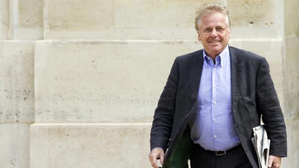 Ein lächelnder Mann mit Brille und Anzug steht vor einer hellen Wand.