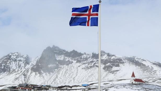 Die isländische Flagge weht vor einer schneebedeckten Landschaft mit Bergen und einer kleinen Kirche.