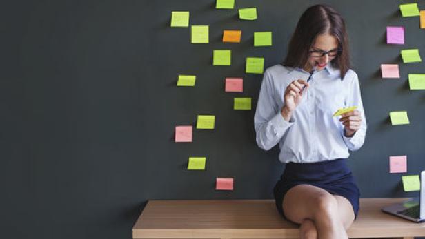 Eine Frau mit Brille sitzt auf einem Tisch vor einer Wand voller Notizen und hält einen Stift.