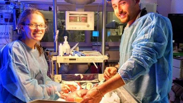 Zwei Personen in OP-Kleidung kümmern sich um ein Neugeborenes in einem Inkubator.