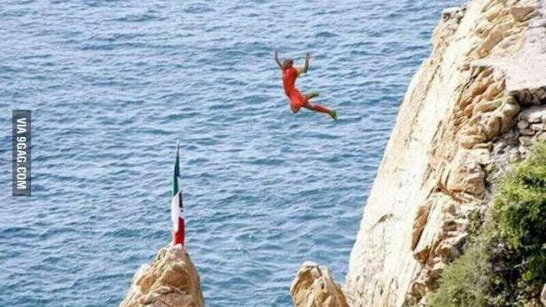 Ein Klippenspringer springt in roter Kleidung ins Meer, im Hintergrund eine mexikanische Flagge.