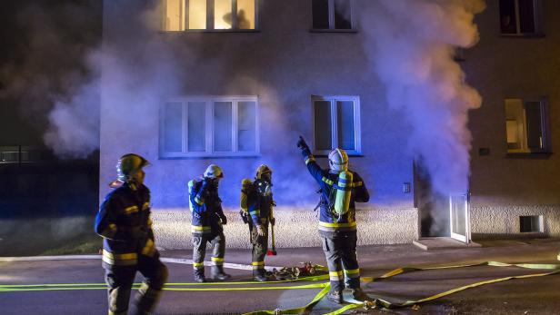 Feuerwehrleute stehen vor einem Gebäude, aus dessen Tür Rauch dringt.