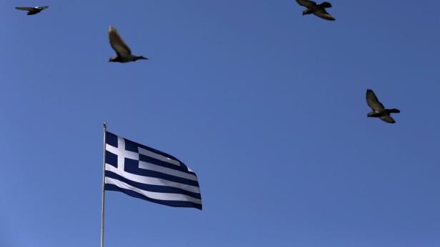 Die griechische Flagge weht vor blauem Himmel, während Vögel fliegen.