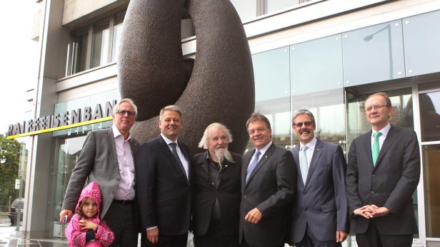 Eine Gruppe von Personen posiert vor einer Skulptur und einem Gebäude der Raiffeisenbank.
