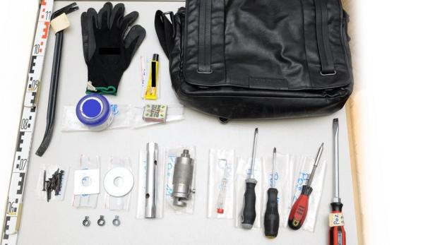 Ein Brecheisen, Handschuhe, Schraubenzieher und andere Werkzeuge liegen neben einer schwarzen Tasche.