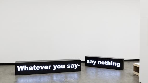 Zwei schwarze Kästen mit Leuchtschrift zeigen die Worte „Whatever you say-“ und „say nothing“.