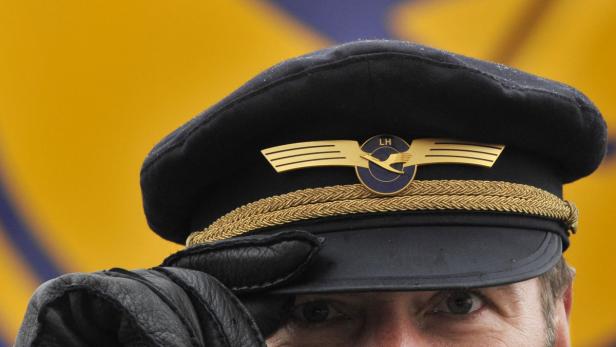 Ein Lufthansa-Pilot mit Mütze und Lederhandschuhen vor gelbem Hintergrund.