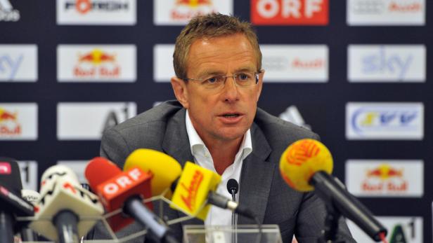 Ein Mann mit Brille spricht vor einer Wand mit Logos in mehrere Mikrofone.