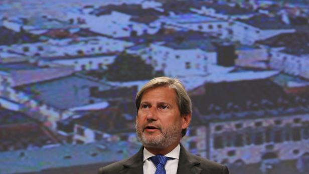 Ein Mann mit Bart und Anzug steht vor einem unscharfen Hintergrund mit Häusern.