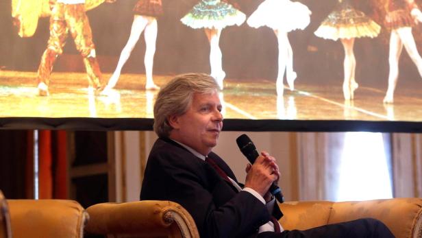 Ein Mann sitzt auf einem Sofa und spricht in ein Mikrofon; im Hintergrund eine Ballettvorführung.