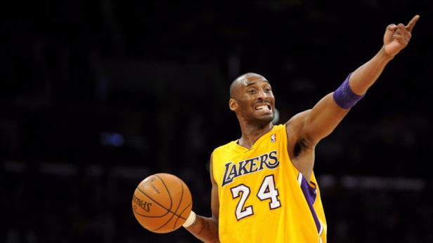 Kobe Bryant im gelben Lakers-Trikot mit der Nummer 24 hält einen Basketball und zeigt nach oben.