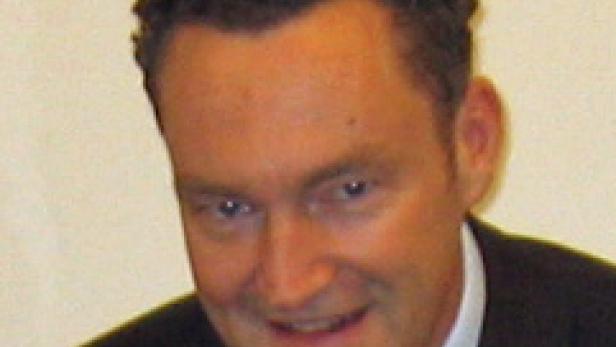Ein Mann mit dunklem, lockigem Haar und einem Anzug lächelt in die Kamera.