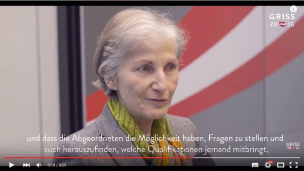 Eine ältere Frau mit kurzem, grauem Haar spricht vor einem Hintergrund mit österreichischen Farben.