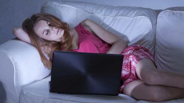 Eine junge Frau schläft auf einem Sofa vor ihrem Laptop ein.