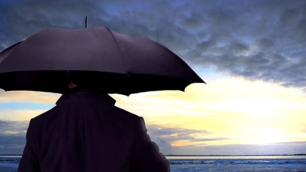 Ein Mann mit dunklem Anzug und Schirm blickt auf das Meer im Abendlicht.