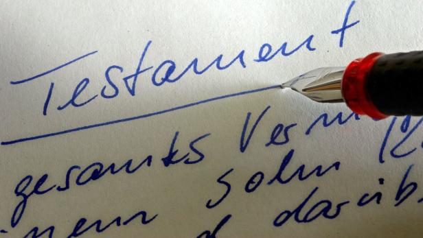 Eine Feder schreibt das Wort „Testament“ auf ein Blatt Papier.