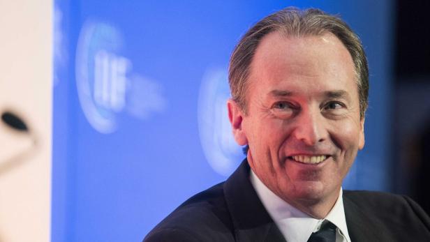 Ein lächelnder Mann im Anzug vor einem blauen Hintergrund mit einem Logo.