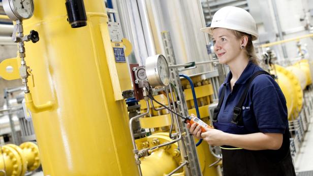 Eine Frau in Arbeitskleidung und Schutzhelm inspiziert eine gelbe Industrieanlage.