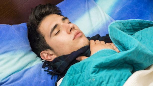 Ein junger Mann schläft mit einem Schal im Bett unter einer türkisfarbenen Decke.