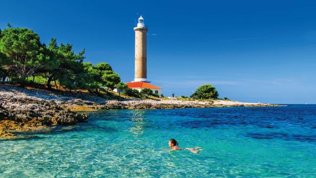 Eine Frau schwimmt im türkisfarbenen Wasser vor einem Leuchtturm an der Küste.