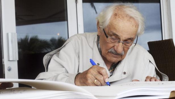 Ein älterer Mann mit Brille und Schnurrbart schreibt mit einem blauen Stift in ein Buch.