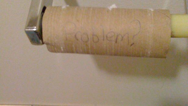 Eine leere Toilettenpapierrolle hängt am Halter, eine weitere liegt auf dem Boden. Auf der Rolle steht „Problem?".