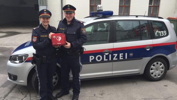 Zwei Polizisten stehen vor einem Polizeiauto und halten einen Defibrillator.