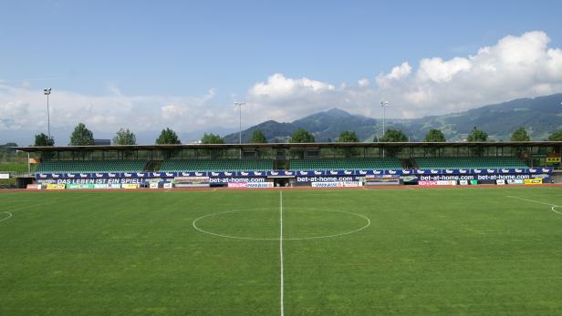 Ein Fußballstadion mit grüner Tribüne und Blick auf bewaldete Hügel.