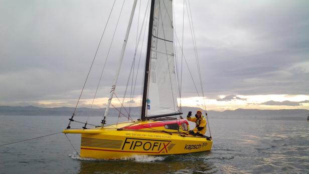 Ein Segler winkt von einem gelben Segelboot mit der Aufschrift „Fipofix“ auf dem Wasser.