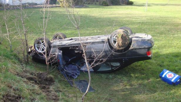 Ein schwarzes Auto liegt auf dem Dach in einem Graben, neben einem Erste-Hilfe-Kasten.