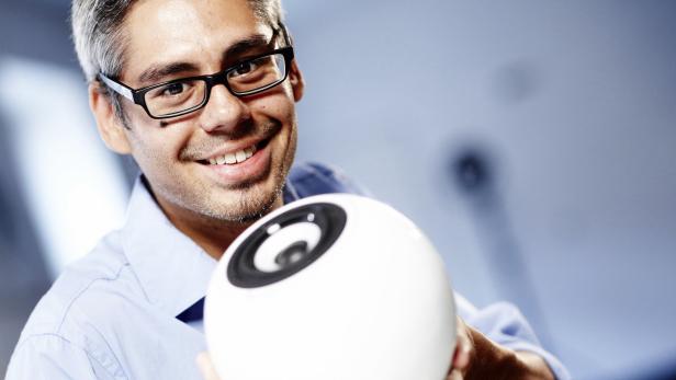 Ein lächelnder Mann mit Brille hält einen runden, weißen Lautsprecher in der Hand.