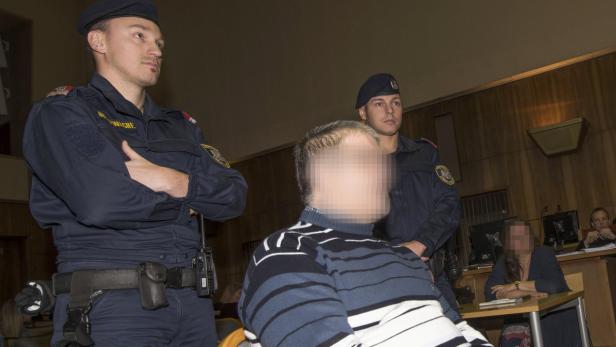 Ein Mann mit verpixeltem Gesicht wird von zwei Polizisten in einem Gerichtssaal bewacht.