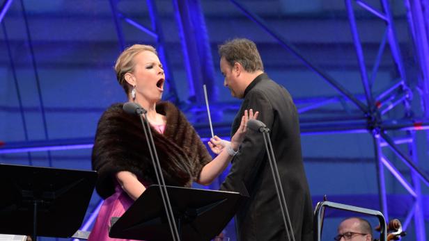 Eine Opernsängerin singt mit einem Dirigenten vor einem Orchester.