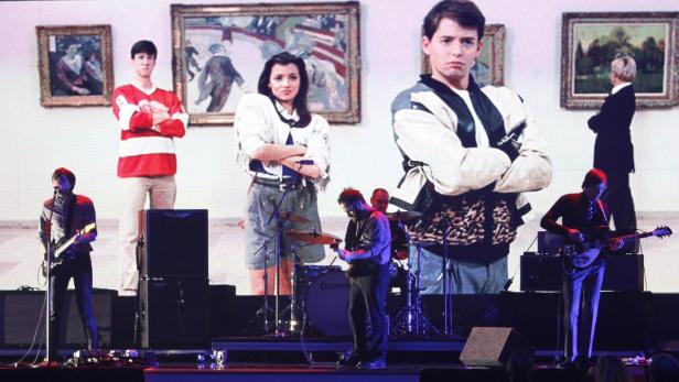 Eine Band spielt vor einer Leinwand mit Szenen aus dem Film „Ferris macht blau“.