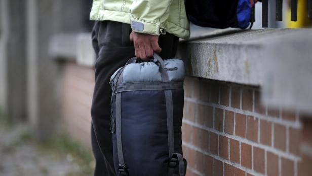 Eine Person hält einen Schlafsack vor einer Backsteinmauer.