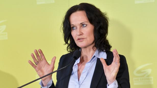 Eine Frau mit dunklen Haaren gestikuliert vor einem grünen Hintergrund mit dem Logo der Grünen.