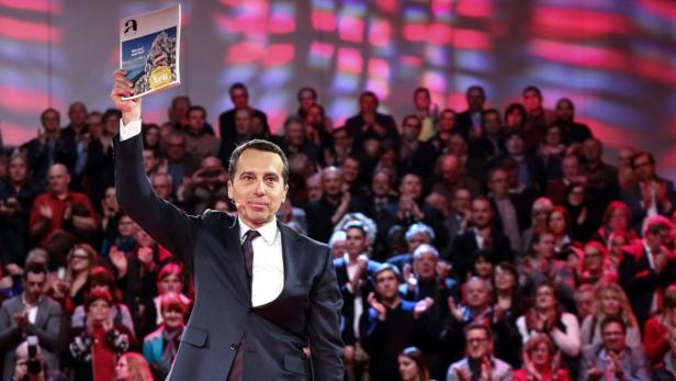 Ein Mann im Anzug hält eine Broschüre vor einer applaudierenden Menschenmenge hoch.