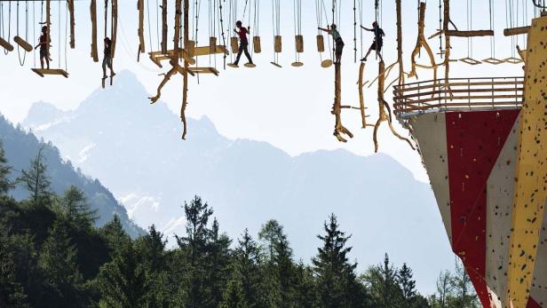 Personen überqueren einen Hochseilgarten vor einer Bergkulisse.