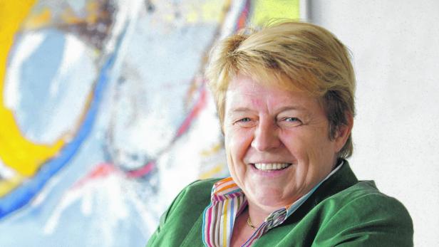 Eine lächelnde Frau mit blonden Haaren und grünem Blazer vor einem abstrakten Gemälde.