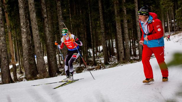 Eine Biathletin läuft auf Skiern durch den verschneiten Wald, während ein Trainer sie begleitet.