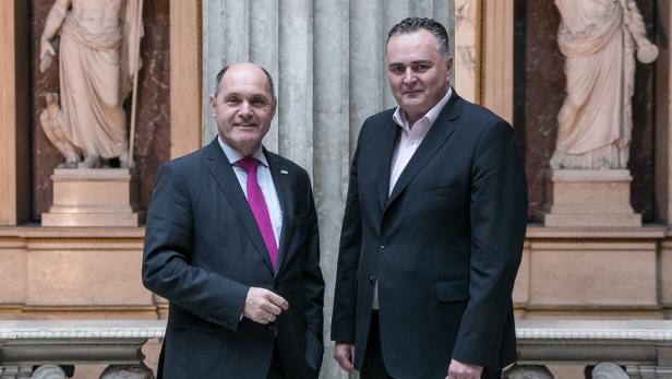 Zwei Männer in Anzügen posieren vor Statuen in einem Gebäude.