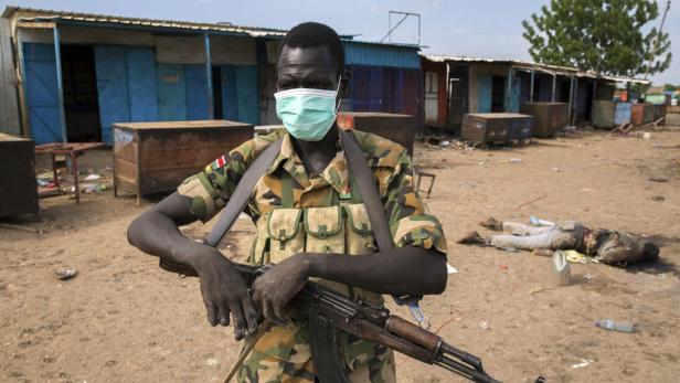Ein Soldat mit Maske und Gewehr steht vor einem zerstörten Gebäude, im Hintergrund eine Leiche.