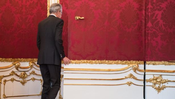 Ein Mann in Anzug steht vor einer roten Wand mit goldenen Verzierungen.